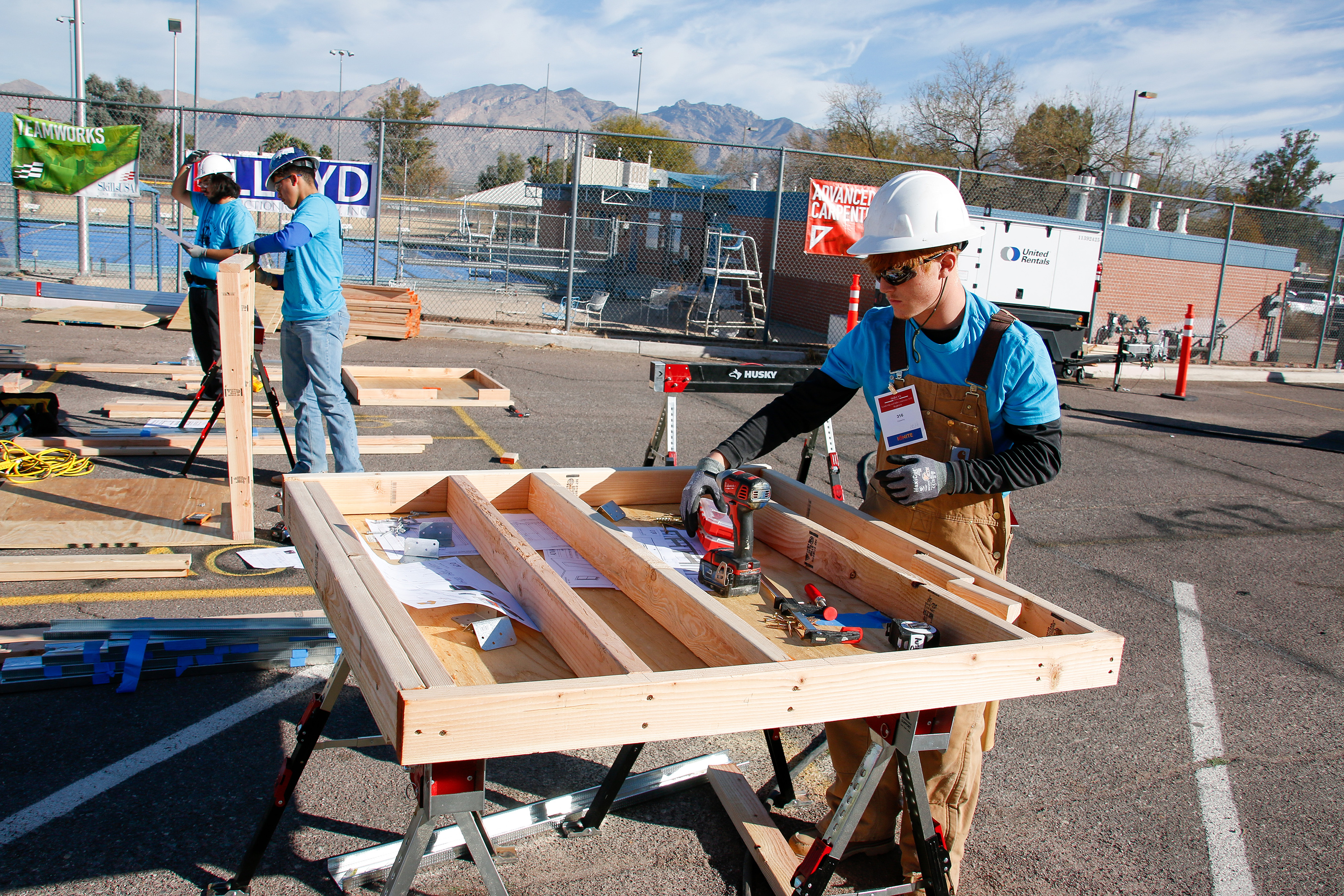 A boy in a white hard hat and overalls holds a drill as he works on a wooden fixture