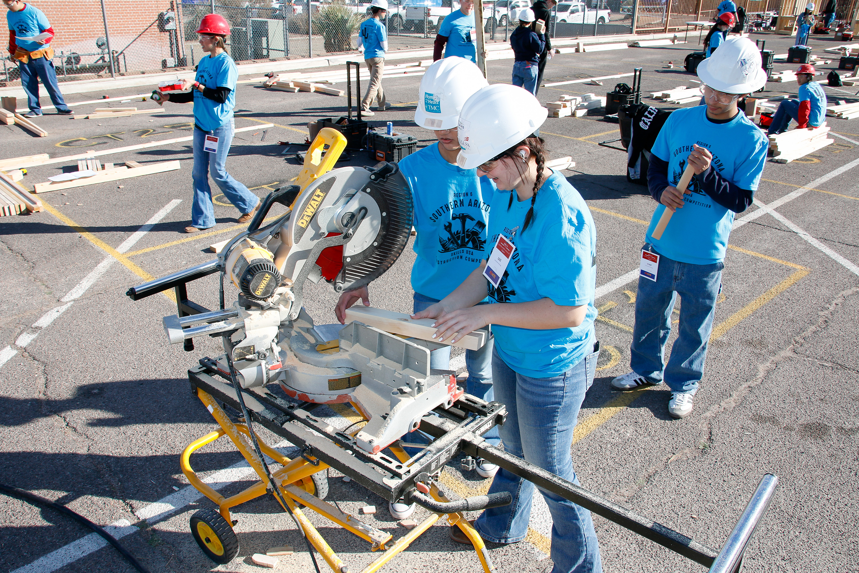 Two girls in white hard hats work on cutting a piece of wood with a machine