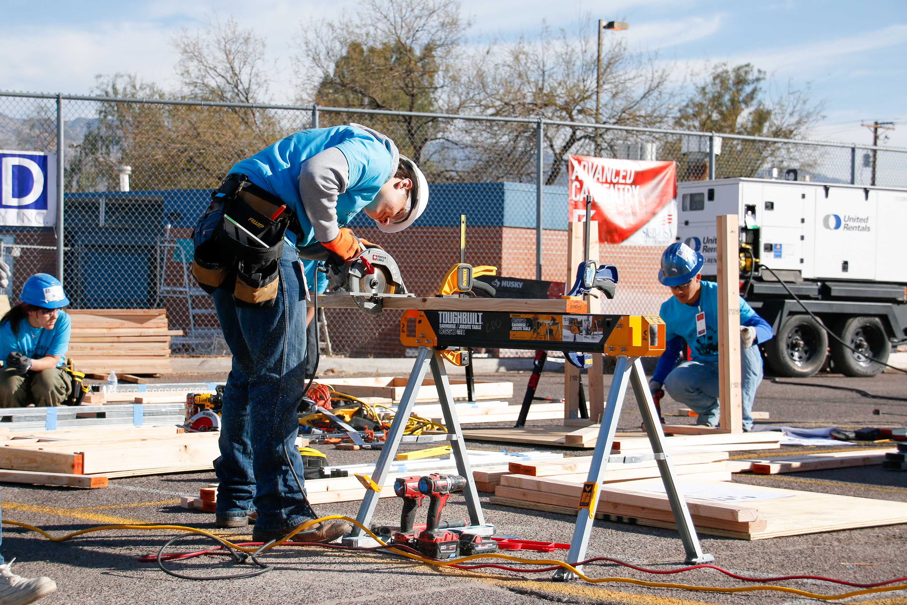 a boy in a white hard hat leans over as he saws a piece of wood