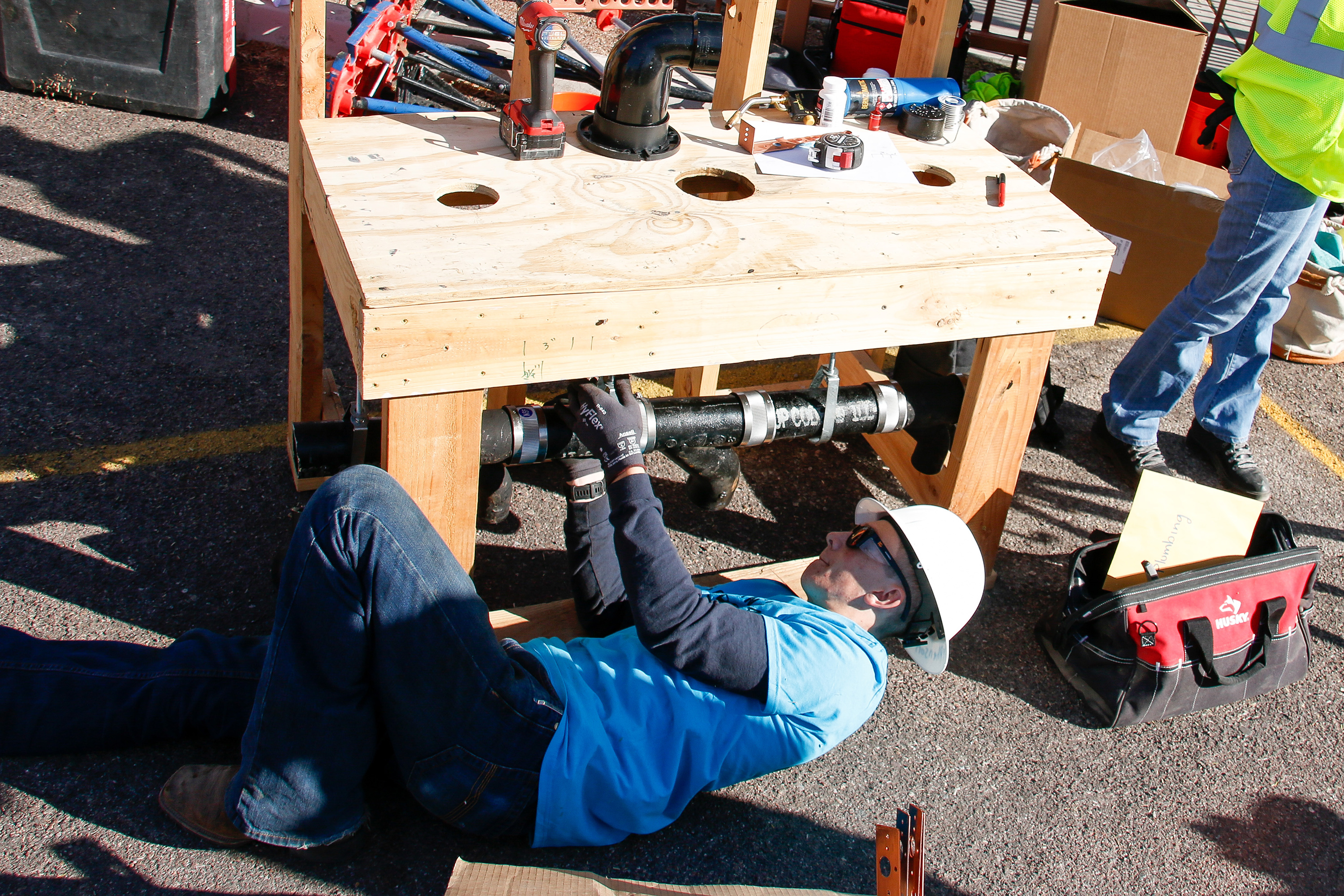 A boy in a white hard hat lays on the ground to fix a black pipe