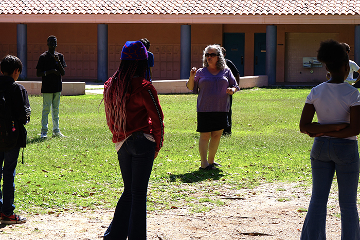 Students stand outside on the field listening to a teacher