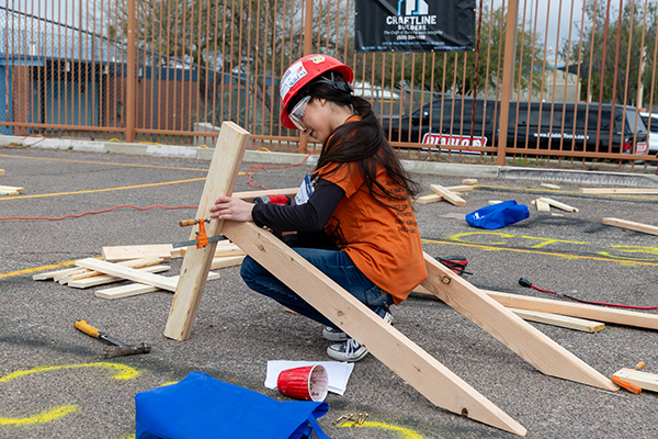 A girl in a hard hat and safety goggles nails boards together
