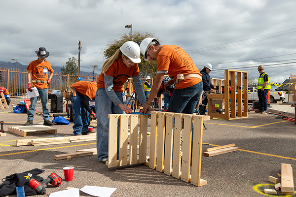 Two students in hard hats and safety goggles nail together some boards