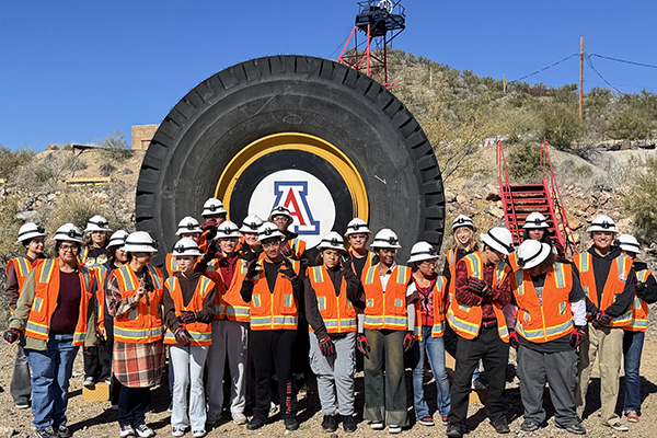 Students wearing hard hats and orange safety vests stand in front of a giant tire outside the mine