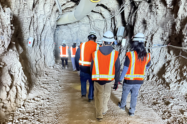 Students wearing hard hats and orange safety vests walk into the mine