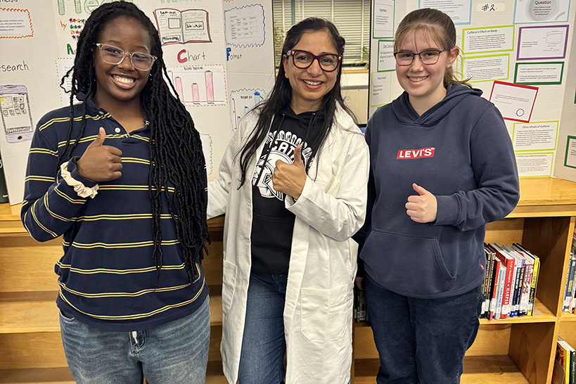 Three teen girls give a thumbs up in front of science fair displays
